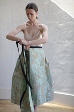 Load image into Gallery viewer, A person with long brown hair, tied back, is standing in a bright room. They are wearing a beige sleeveless top and a green floral-patterned skirt. The person is holding a black belt or ribbon and seems to be in the process of tying it around their waist, accentuating their handmade, limited edition Dupioni Pleated Wrap Skirt | Jade attire.