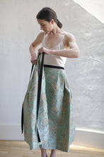 Load image into Gallery viewer, A person with long dark hair in a ponytail stands against a light-colored wall, wearing a sleeveless top and holding a Dupioni Pleated Wrap Skirt | Jade made of handmade Dupioni silk with a floral pattern and black waistband, in the process of either putting it on or taking it off.