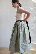Load image into Gallery viewer, A person with dark hair, tied back, facing to the left and adjusting their sleeveless beige top with a long black ribbon. They are wearing a Dupioni Pleated Wrap Skirt | Jade made of Limited Edition Dupioni silk featuring an intricate floral pattern. The background is a plain white wall with wooden flooring.