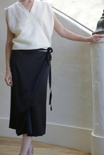 Load image into Gallery viewer, A person in a sleeveless white blouse and the elegant Charmeuse Petal Wrap Skirt in black, featuring a cashmere waistband and zero-waste design, stands by a curved white railing with one hand resting on it. The softly lit, neutral background enhances the outfit's simple elegance.