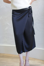 Load image into Gallery viewer, A person in a white top wears the Charmeuse Petal Wrap Skirt in black, which features a tied bow on the side. The neutral background includes a light-colored wall and wooden floor, and their feet are in light-hued open-toe shoes.