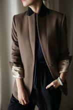 Load image into Gallery viewer, Sloane is showcased in a dark brown coat featuring structured shoulders, with the sleeves stylishly rolled up to reveal a light-colored lining. Paired with dark pants and a bracelet adorning the left wrist, the ensemble exudes sophistication. The individual strikes a casual pose, standing with one hand in their pocket, though their face remains out of view.