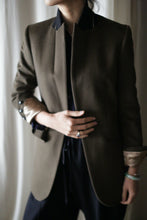 Load image into Gallery viewer, A person is dressed in the "Sloane" blazer, featuring a dark green color with structured shoulders and black and beige accents. Their left hand, adorned with a ring and bracelet, rests on their midsection. The light-colored curtain serves as the background, while the person's face remains unseen in the image.