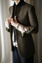 Load image into Gallery viewer, Someone wearing the "Sloane" blazer with its dark, tailored design and structured shoulders over a white top and dark pants adjusts their collar. The background is softly blurred, accentuating the textures and details of the clothing. The lighting is soft and warm, creating a stylish and sophisticated atmosphere.