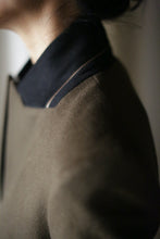 Load image into Gallery viewer, Close-up of a person wearing the Sloane, a dark-colored, woven cashmere coat with a black collar. The image highlights the neckline and structured shoulders of the garment, showcasing the texture and details of the material, while the background is softly blurred.