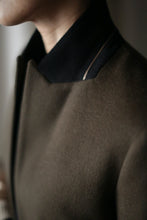 Load image into Gallery viewer, Close-up of a person wearing the Sloane coat in dark green or olive, accented by a black collar. The image captures only the lower part of the face and the top portion of this equestrian-length coat, highlighting the rich textures and colors of its woven cashmere fabric. The background is blurred.