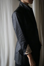 Load image into Gallery viewer, A person is wearing a dark-colored Waxed Linen Sloane tuxedo jacket with the sleeves rolled up to reveal a beige lining. The shot captures the person from neck to mid-thigh, with their head slightly turned away from the camera. The background is softly lit, featuring light-colored curtains.