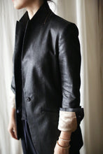 Load image into Gallery viewer, A person wearing the Waxed Linen Sloane blazer with the sleeves rolled up, revealing a light-colored inner lining. The individual, whose head is turned away, stands against a light-colored curtain backdrop, sporting subtle wrist jewelry to complete the outfit.