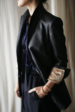 Load image into Gallery viewer, A person dressed in a Waxed Linen Sloane tuxedo jacket with rolled-up sleeves and navy blue clothing underneath stands with one hand in their pocket. Their head is slightly turned away from the camera, and the background features a light, draped fabric.