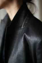 Load image into Gallery viewer, Close-up of a person wearing the Waxed Linen Sloane jacket in black, featuring a high collar. The focus is on the shoulder and upper chest area, highlighting the fabric's sheen and texture. The background is out-of-focus, with the person's face partially out of the frame.