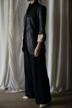 Load image into Gallery viewer, A person stands against a background of soft, light-colored drapery. They are dressed in a Sloane tuxedo jacket with rolled-up sleeves, wide-legged dark trousers, and light-colored shoes. Only part of their face is visible, and they are wearing a Waxed Linen Sloane bracelet on one wrist.