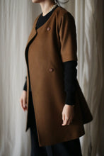 Load image into Gallery viewer, Wearing the Wexley, a brown short-sleeved double-breasted coat with an A-line cut and two visible buttons, over a black long-sleeve top. The softly lit background features white curtains.
