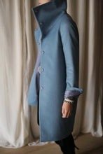 Load image into Gallery viewer, A person wearing the Stamford Long coat, featuring a high-collar design and button details in a beautiful blue shade, stands indoors against a light curtain backdrop. The cashmere fabric of the coat adds an elegant touch, while their right sleeve is partially rolled up to reveal a bracelet on their wrist.