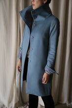 Load image into Gallery viewer, A person wearing the Stamford Long, a stylish high-collared blue cashmere coat, stands against a light curtain background. The coat is buttoned, and the person appears to have their hands in the pockets.