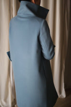 Load image into Gallery viewer, A person wrapped in a high-collared Stamford Long cashmere coat in blue stands with their back to the camera, next to white curtains.