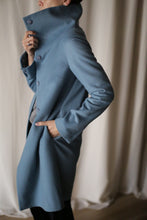 Load image into Gallery viewer, Person wearing the Stamford Long, a stylish high-collared cashmere blue coat, with their hand in the pocket, standing against a softly blurred light-colored curtain background.