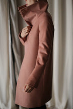 Load image into Gallery viewer, Dressed in a Stamford Long coat with an upturned dramatic collar, a person stands against a curtain backdrop. The individual highlights the coat's details and exquisite fit, crafted from 100% woven cashmere, by placing their hand near the collar.