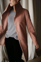 Load image into Gallery viewer, A person wearing a Stamford Long coat with a dramatic collar, crafted from 100% woven cashmere, stands indoors. The high-collared, buttoned-up pink coat elegantly contrasts with the gray sweater and black pants, while cream-colored curtains provide a soft backdrop.