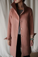 Load image into Gallery viewer, A person wearing the Stamford Long, a pink woven cashmere coat with a high collar turned up over a gray top and black pants. The coat is fastened with three visible buttons. They are accessorized with bracelets on one wrist and a gold necklace. In the background, there is a beige curtain.