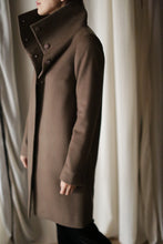 Load image into Gallery viewer, A person stands facing left, wrapped in the Stamford Long coat, a long brown piece crafted from cashmere with a high collar and buttons. The background consists of softly draped, light-colored curtains.