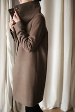 Load image into Gallery viewer, A person wearing the Stamford Long cashmere coat stands with their side facing the camera, gently holding the collar up with one hand. Light curtains create a soft backdrop.
