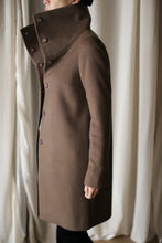 Load image into Gallery viewer, A person wearing the Stamford Long, a high-collared, buttoned brown cashmere coat, stands sideways against a backdrop of light-colored curtains.