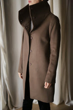Load image into Gallery viewer, A person wearing the Stamford Long, a long brown cashmere coat with a high collar and five buttons, stands indoors facing left. The background consists of softly draped, light-colored curtains.
