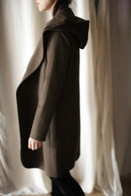 Load image into Gallery viewer, Wearing the Mitchley Long dark brown coat with a hood and handmade snap buttons, a person stands in profile against white curtains. Their face is partially visible, long dark hair falls freely, and the soft lighting creates a warm, fairytale-like shadow.
