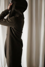 Load image into Gallery viewer, A person wearing the Mitchley Long coat with handmade snap buttons is partially facing away, pulling the hood over their head. Light-colored curtains form the backdrop, creating a subdued mood focused on the act of putting on the hood.