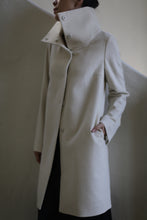 Load image into Gallery viewer, Dressed in the Stamford Long | Undyed, a tailored light beige cashmere coat with a high collar, the person stands against a minimalist neutral backdrop. The coat's button row and hand-in-pocket pose exude modern sophistication.
