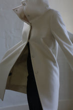 Load image into Gallery viewer, A person wears a Stamford Long | Undyed coat in cashmere featuring a high collar and large buttons. The coat is slightly open, exposing a dark outfit beneath, set against a plain, neutral-toned wall.