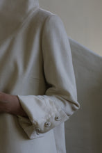 Load image into Gallery viewer, Close-up of a person wearing the Stamford II | Undyed coat with their arm partially visible. The high collar coat features cuffs adorned with neatly folded fabric and three decorative, shiny metallic buttons. The background is softly lit and out of focus.