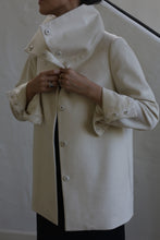 Load image into Gallery viewer, A person wearing the Stamford II | Undyed, a high-collared coat crafted from woven cashmere, adjusts the button-secured cuffs. The scene is set against a simple, light-colored wall and staircase, with the individual's face not visible.