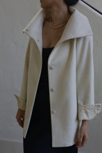 Load image into Gallery viewer, A person is wearing the Stamford II model, a woven cashmere coat in its undyed cream color, featuring large buttons and turned-up cuffs over a black garment. The high-collar coat is complemented by a gold necklace. The individual's head is turned to the side against a plain, light-colored wall.