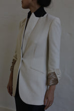 Load image into Gallery viewer, A person wearing the Sloane cashmere undyed blazer, tailored in a cream color with metallic sleeve cuffs, stands in profile against a plain backdrop. The blazer features a sleek design reminiscent of men's tailoring, complete with a concealed front closure and a side pocket.