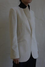 Load image into Gallery viewer, A person dressed in the Sloane cashmere undyed blazer stands against a neutral background, with their face partially out of view. The cream-colored jacket features a black collar reminiscent of a tuxedo, and it is paired with black skirt or pants. The blazer's design showcases simple, elegant lines that reflect the finesse of men's tailoring.
