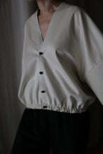 Load image into Gallery viewer, A person wears the Blanc de Chine, a handmade loose-fitting white cashmere top crafted in San Francisco. Featuring a deep V-neckline, long sleeves, black buttons, and an elastic waistband, it's styled with dark pants. The scene is softly lit against a dark background.