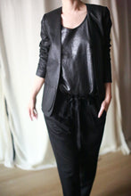 Load image into Gallery viewer, A person wearing a Waxed Linen Blazer over a shiny black top and black pants, with one hand in their pocket. The background features white curtains.