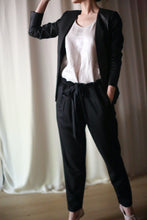 Load image into Gallery viewer, A person stands indoors in front of a light-colored curtain, dressed in a black Waxed Linen Blazer, a white top, and black drawstring waist pants. Their right hand is raised near their head while their left hand rests in their pocket. Their face is not visible.