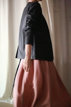 Load image into Gallery viewer, A person is partially visible from the head down, wearing a black Waxed Linen Blazer and a flowing pink skirt. The individual stands in soft lighting against a neutral backdrop. The pose is relaxed, and the clothing creates a striking contrast in texture and color.