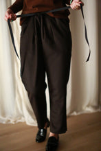 Load image into Gallery viewer, A person is shown tying a silk ribbon drawstring on Cashmere Unisex Pants | Brown. The background features soft white curtains, and the person is wearing a brown top and black shoes. Only the lower half of the body is visible, highlighting the unisex appeal of the outfit.