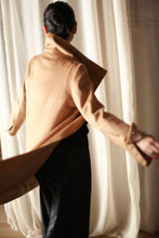 Load image into Gallery viewer, A person with dark hair in a bun is seen from the back, wearing the Stamford Long tan cashmere overcoat that flows as they move. The coat's collar stands elegantly, complementing their black pants as they stand in front of sheer white curtains.