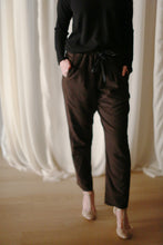 Load image into Gallery viewer, A person wearing a black long-sleeve top and Cashmere Unisex Pants | Brown with hands in pockets stands on a wooden floor. The background features sheer white curtains. Only the person's torso and legs are visible, giving a minimalist, unisex appeal to the outfit.