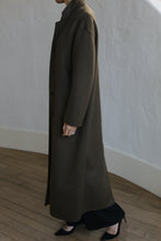 Load image into Gallery viewer, Dressed in the Bamford Long Double-face Cashmere coat in Olive, a person stands stylishly indoors against a light-textured wall. Viewed from the side, their ring-adorned hand adds elegance to the scene, complemented by the wooden floor beneath.