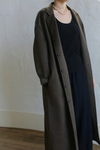 Load image into Gallery viewer, A person stands against a light wall, wearing an olive Bamford Long Double-face Cashmere coat. The luxurious coat, partially buttoned, overlays a black top and trousers. A simple necklace adds elegance, while the image emphasizes the clothing over facial details.