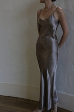 Load image into Gallery viewer, A woman stands against a light-colored wall wearing the Liquid Slip Dress Long | Mink made of plant-derived material. The silver satin slip dress features thin straps and a bias cut, creating a fitted silhouette that flares slightly at the bottom. She also wears a delicate gold necklace, with her hands tucked behind her back.