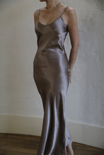 Load image into Gallery viewer, A person is wearing a Liquid Slip Dress Long | Mink made from plant-derived material, featuring a satin, silvery-gray slip design with thin straps. They are also wearing a thin gold necklace. The background is plain with light-colored walls, and the floor is wooden. The image is taken indoors with soft lighting.