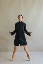 Load image into Gallery viewer, A ballet dancer gracefully performs a balanced pose on pointe, wearing the Edmonton black coat with a reversible collar. She stands against a neutral background, arms gently outstretched and legs crossed, embodying elegance and strength.