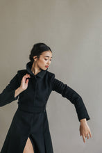 Load image into Gallery viewer, A person in a Hyde black, woven cashmere coat gracefully poses against a plain, light-colored background. They have dark hair pulled back and an elegant expression, with one arm raised and the other lowered, conveying a sense of movement.