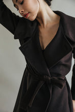 Load image into Gallery viewer, A person with dark hair poses in the Malvern, a luxurious double-breasted coat, with one arm raised. The neutral background and downward gaze create a contemplative atmosphere, highlighting the sophisticated elegance of the woven cashmere fabric.